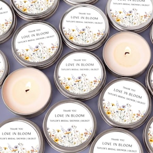 May include: A collection of round silver metal candle tins, some with lids featuring a floral print and the text "THANK YOU LOVE IN BLOOM TAYLOR'S BRIDAL SHOWER | 08.29.27", and others open to reveal a cream-colored wax candle with a lit wick.