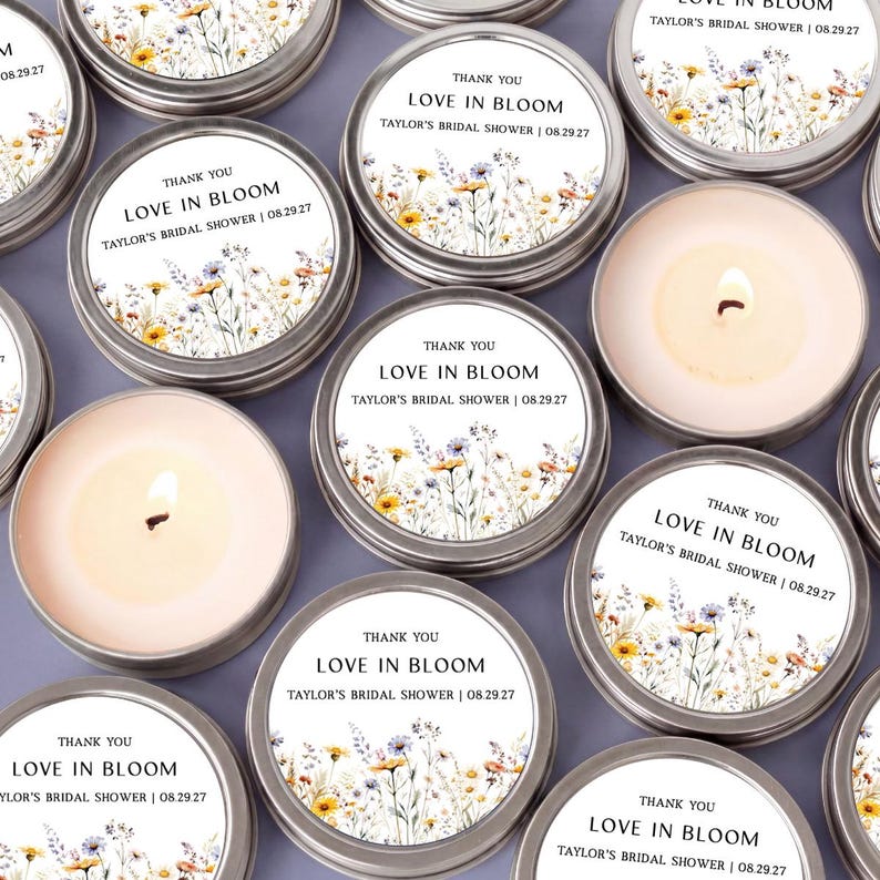 May include: A collection of round silver metal candle tins, some with lids featuring a floral print and the text "THANK YOU LOVE IN BLOOM TAYLOR'S BRIDAL SHOWER | 08.29.27", and others open to reveal a cream-colored wax candle with a lit wick.