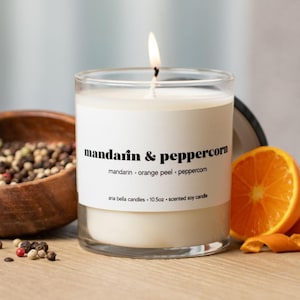 May include: A lit scented soy candle in a clear glass jar with a white label that reads "mandarin & peppercorn." The candle is next to a wooden bowl of peppercorns and a sliced orange. The candle is 10.5 oz.