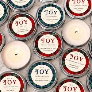 May include: A collection of round metal candle tins featuring lids with red or blue plaid patterns and circular labels. The labels display the text IT'S A JOY followed by personalized messages such as TO STYLE YOU, TO WORK WITH YOU, or TO TRAIN WITH YOU, along with sender names.