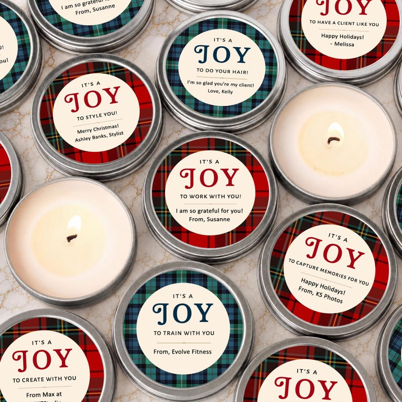 May include: A collection of round metal candle tins featuring lids with red or blue plaid patterns and circular labels. The labels display the text IT'S A JOY followed by personalized messages such as TO STYLE YOU, TO WORK WITH YOU, or TO TRAIN WITH YOU, along with sender names.