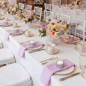 May include: A formal table setting featuring a white tablecloth, light purple cloth napkins, silver metal tins with circular labels, white ceramic teacups on saucers, silver cutlery, and floral centerpieces with pastel flowers in white vases.
