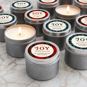 May include: A collection of round silver metal candle tins with lids featuring circular labels. The labels display various plaid patterns in red, green, and blue, with text including "IT'S A JOY" and personalized messages from senders.