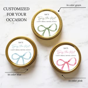 May include: Three round gold metal tins with white labels featuring a watercolor bow illustration. Each label displays the text "SHE'S Tying the Knot" and "CASEY'S BRIDAL SHOWER | 05.05.27" in green, blue, or pink ink, matching the color of the bow.