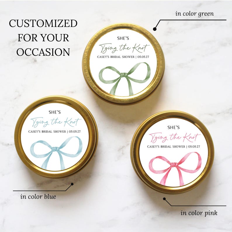 May include: Three round gold metal tins with white labels featuring a watercolor bow illustration. Each label displays the text "SHE'S Tying the Knot" and "CASEY'S BRIDAL SHOWER | 05.05.27" in green, blue, or pink ink, matching the color of the bow.