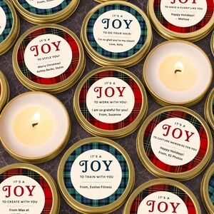 May include: A collection of round gold metal candle tins featuring circular labels with red or blue plaid patterns. Each label displays the text IT'S A JOY followed by personalized messages such as TO STYLE YOU, TO DO YOUR HAIR, or TO WORK WITH YOU, including sender names.
