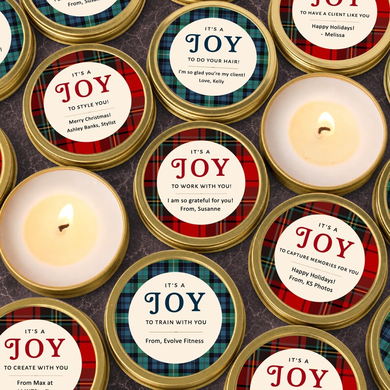 May include: A collection of round gold metal candle tins featuring circular labels with red or blue plaid patterns. Each label displays the text IT'S A JOY followed by personalized messages such as TO STYLE YOU, TO DO YOUR HAIR, or TO WORK WITH YOU, including sender names.