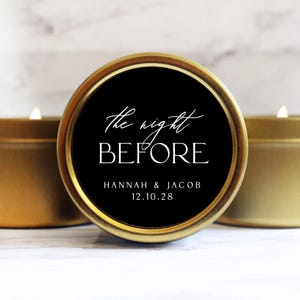 May include: A round gold metal candle tin with a black circular label featuring the white text The night BEFORE, followed by the names HANNAH & JACOB and the date 12.10.28. The candle is lit, and the lid is positioned to show the label clearly.