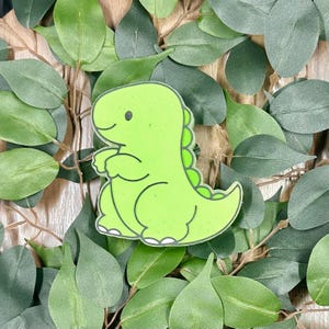 May include: A lime green dinosaur-shaped sticker with a black outline and white accents. The dinosaur has a friendly expression and a row of green spikes down its back. The sticker is set against a backdrop of green leaves.