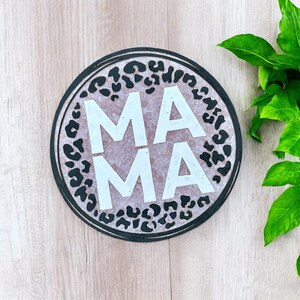 May include: A round wooden sign with a black and white leopard print design. The word "MAMA" is written in white letters on the sign.