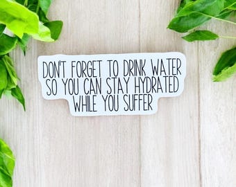 May include: A white sticker with black text that reads "DON'T FORGET TO DRINK WATER SO YOU CAN STAY HYDRATED WHILE YOU SUFFER." The sticker is surrounded by green leaves and rests on a light wood surface.