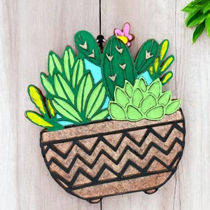 May include: A wooden cutout of a potted succulent plant with a brown pot with a chevron pattern. The succulent is green and has a pink flower.