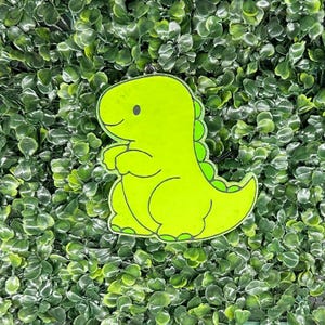 May include: A neon green dinosaur-shaped keychain. The cartoon dinosaur has a smiling face, small arms, and a row of green spikes down its back. The keychain is set against a backdrop of green foliage.