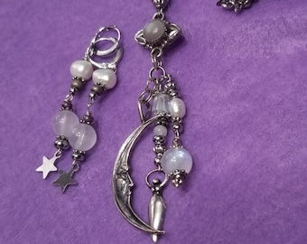 May include: A silver crescent moon charm with a face and a small figure standing on the crescent. The charm is attached to a silver chain with white and gray beads. The chain also has two silver stars.