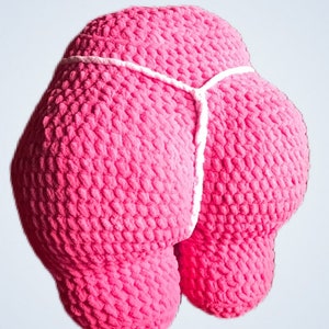 Peut inclure: Un coussin rose en crochet en forme de fesses, avec un d&eacute;tail de ficelle blanche. Le texte "Booty Pillow" est affich&eacute; dans un rectangle rose en haut de l'image. L'oreiller est un article fantaisie.