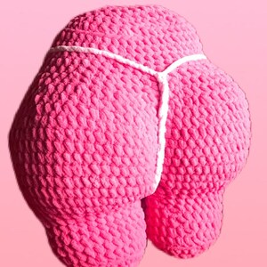 Peut inclure: Un coussin rose en crochet en forme de fesses, avec un d&eacute;tail de ficelle blanche. Le texte "Booty Pillow" est affich&eacute; dans un rectangle rose en haut de l'image. Le fond est rose uni.