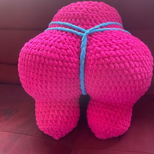 Peut inclure: Un grand coussin rose en crochet, en forme de silhouette stylis&eacute;e. Un cordon bleu clair en crochet entoure le milieu du coussin. Le coussin est pos&eacute; sur une surface rouge.