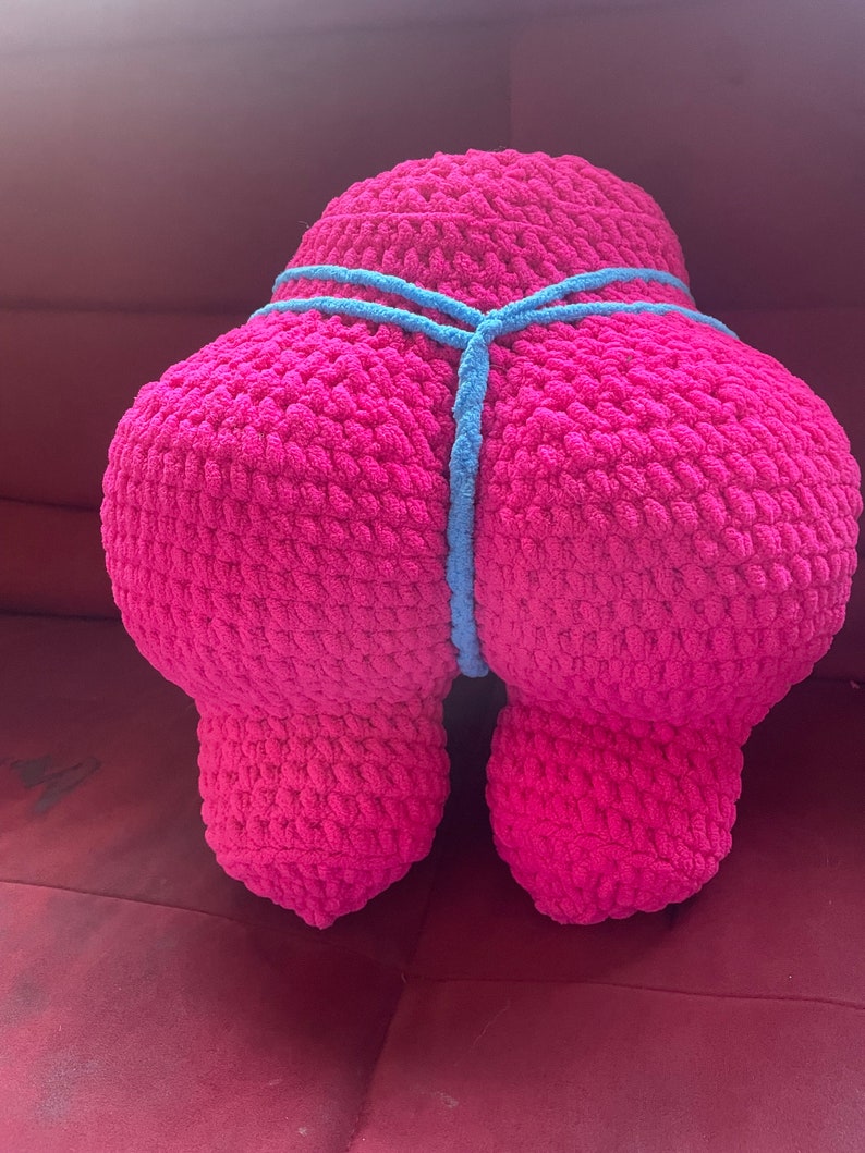 Peut inclure: Un grand coussin rose en crochet, en forme de silhouette stylis&eacute;e. Un cordon bleu clair en crochet entoure le milieu du coussin. Le coussin est pos&eacute; sur une surface rouge.