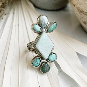 May include: A silver ring with a turquoise and white gemstone design. The ring features a large, diamond-shaped white gemstone surrounded by smaller turquoise gemstones.