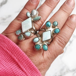 Puede incluir: Dos anillos de plata con piedras turquesa y ópalo. Los anillos están en una mano con una camisa rosa.