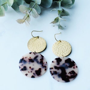 May include: A pair of gold and white statement earrings. The earrings feature a hammered gold disc and a white disc with a tortoiseshell pattern.