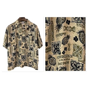 May include: Tan button-down shirt with a repeating pattern of pineapples, flowers, and geometric shapes in black and olive green. The shirt has short sleeves and a collared neckline.