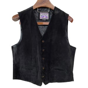 Può includere: Gilet in pelle scamosciata nera con scollo a V e chiusura con bottoni. Il gilet ha due tasche anteriori e un'etichetta con la scritta "WATERSHED". Il gilet è appeso a una gruccia di legno.