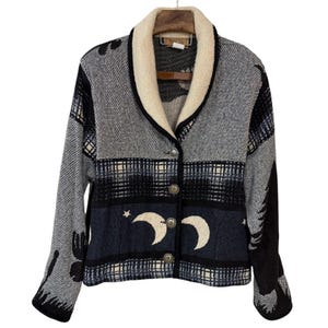May include: A long-sleeve button-up cardigan featuring a cream-colored shawl collar and a woven pattern of black, gray, and navy blue. The design includes crescent moon and star motifs across the lower section, finished with four silver-toned buttons.