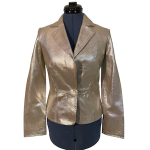 May include: A metallic gold blazer with a notched lapel and a single-button closure. The jacket has long sleeves and a tailored fit, displayed on a black dress form. The blazer's shiny finish catches the light, creating a shimmering effect.
