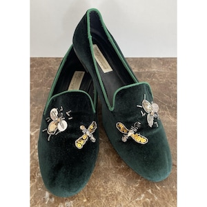 May include: A pair of dark green velvet loafers with green trim. Each shoe features an embroidered bee design with gold sequins and silver beads. The shoes have a classic slip-on style.