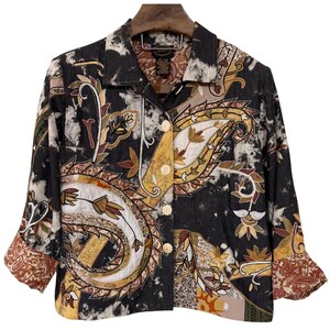 Puede incluir: Una chaqueta de manga larga con botones que presenta una base de teñido anudado en blanco y negro con grandes patrones de cachemira bordados y coloridos en tonos dorados, marrones y crema. La prenda tiene cuello clásico, cuatro botones claros y puños.