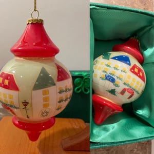 May include: A Christmas ornament with a red top and bottom, and a cream-colored body. The body is decorated with painted houses, trees, and the word "Pier" and "Candy". The ornament is hanging from a gold string.