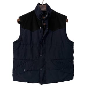 May include: A navy blue quilted vest with a black yoke and collar. The vest features a zip closure, snap pockets, and a stand-up collar. It is suspended on a wooden hanger.