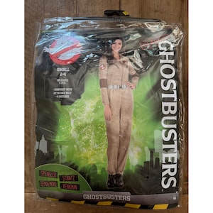 May include: A Ghostbusters costume in a clear plastic package featuring a tan jumpsuit with an attached belt and four patches. The packaging displays the Ghostbusters logo, the text GHOSTBUSTERS, and the names Spengler, Stantz, Zeddemore, and Venkman.