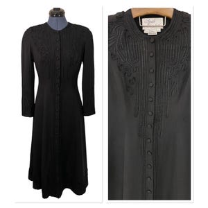May include: A long, black dress with intricate embroidered detailing. The dress features a button-down front, long sleeves, and a tailored silhouette. The embroidery is concentrated around the neckline, down the front, and on the sleeves. The dress is displayed on a mannequin.