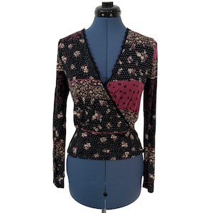 Puede incluir: Una blusa cruzada de manga larga con base negra, lunares blancos y estampados florales en tonos rosa, crema y verde. La prenda tiene un escote en V ribeteado con encaje negro y un diseño tipo patchwork con paneles de tela contrastantes.