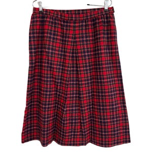 May include: A red plaid skirt with a pleated front, hanging on a hanger. The skirt features a classic tartan pattern in red, navy blue, and gold. The skirt is made of a woven fabric.