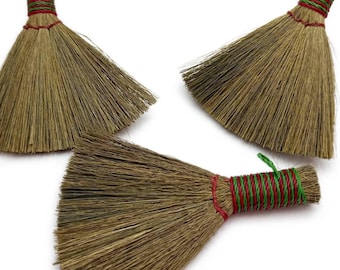 May include: Three small hand brooms made of natural tan straw fibers. Each broom features a handle wrapped in alternating red and green thread, with a small green loop at the top for hanging. The bristles are fanned out in a triangular shape.