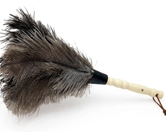 May include: A feather duster featuring long, dark gray ostrich feathers attached to a black ferrule. The handle is light-colored, carved wood with a smooth, ribbed texture and a thin brown leather cord loop attached to the end for hanging.