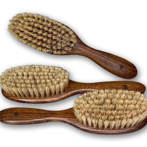 May include: Three oval-shaped hair brushes with dark brown wooden handles and light tan natural bristles. Each brush features a smooth, polished wood grain finish and a small, dark logo mark stamped on the handle near the brush head.