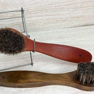 May include: Two wooden shoe brushes with dark bristles. One brush has a reddish-brown handle, and the other has a darker brown handle. Both handles have a small, stylized design. The brushes are displayed on a light-colored wooden surface.