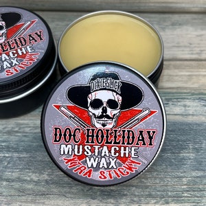 May include: A silver tin with a black and white skull wearing a black cowboy hat and holding two pistols. The tin has red text that reads "Doc Holliday Mustache Wax Extra Sticky" and a smaller black text that reads "Dixie Slick".