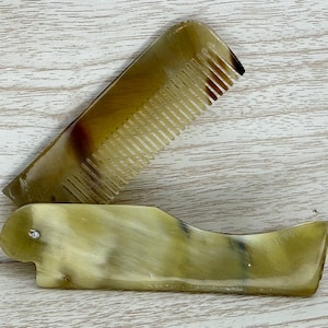 May include: A folding comb made of brown and beige horn. The comb has a long, narrow handle and a row of teeth. The comb is closed and folded in half.