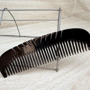 May include: A black comb with a curved handle and evenly spaced teeth. The comb is made of a glossy material and is resting on a light-colored wooden surface. A metal wire structure is in the background.