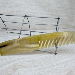 May include: A yellow comb with fine teeth, resting on a wire display stand. The comb has a curved shape and a smooth, polished surface. The background features a wire structure and a light-colored wooden surface.