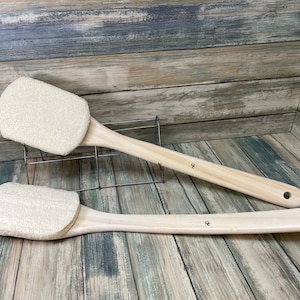 May include: Two wooden bath brushes with natural loofah sponges attached. The brushes have a long, thin handle and are lying on a wooden surface.