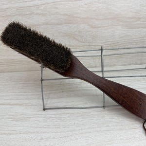 May include: A dark brown shoe brush with a long, curved wooden handle and a dark bristle head. The handle has a leather loop for hanging. The brush is resting on a wire rack.