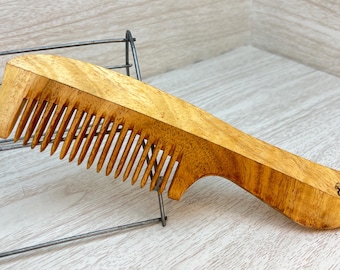 Pode incluir: Um pente de madeira com acabamento marrom natural e cabo curvo. O pente tem um design de dentes largos e um pequeno logotipo no cabo.