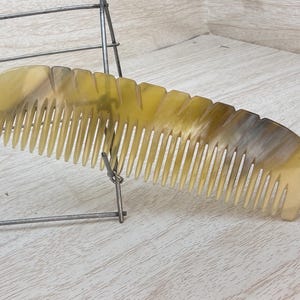 May include: A light-colored comb with a curved handle and closely spaced teeth. The comb is made of a translucent material with variations of yellow and brown. It rests on a light-colored wooden surface.