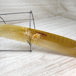 May include: A golden-yellow comb with fine teeth, resting on a light-colored wooden surface. The comb has a smooth, curved design and is made of a translucent material. A wire rack is in the background.